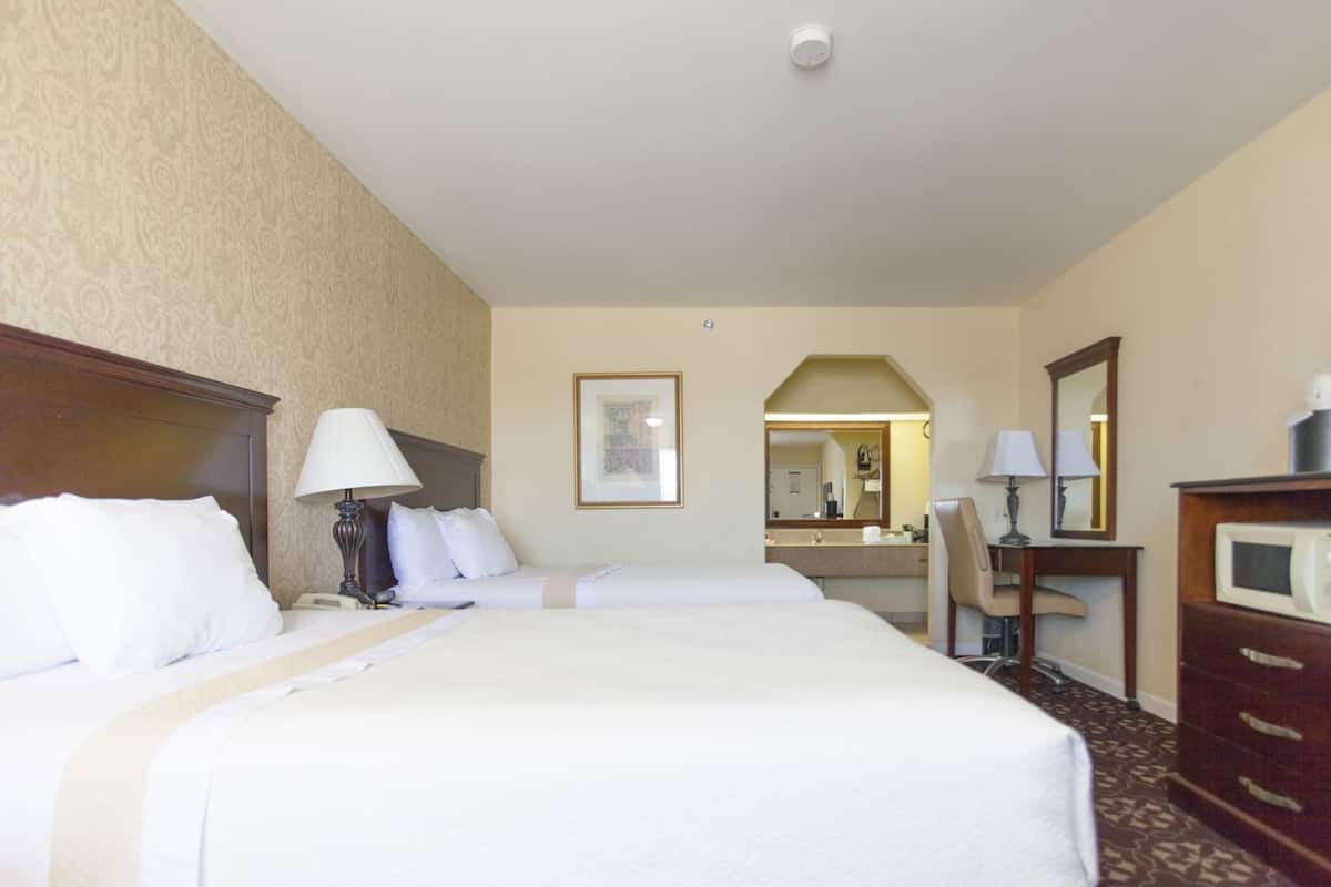 Une chambre d’hôtel avec un grand lit, deux tables de chevet avec des lampes, un bureau avec une chaise, un four à micro-ondes, un miroir et un tableau encadré au mur.