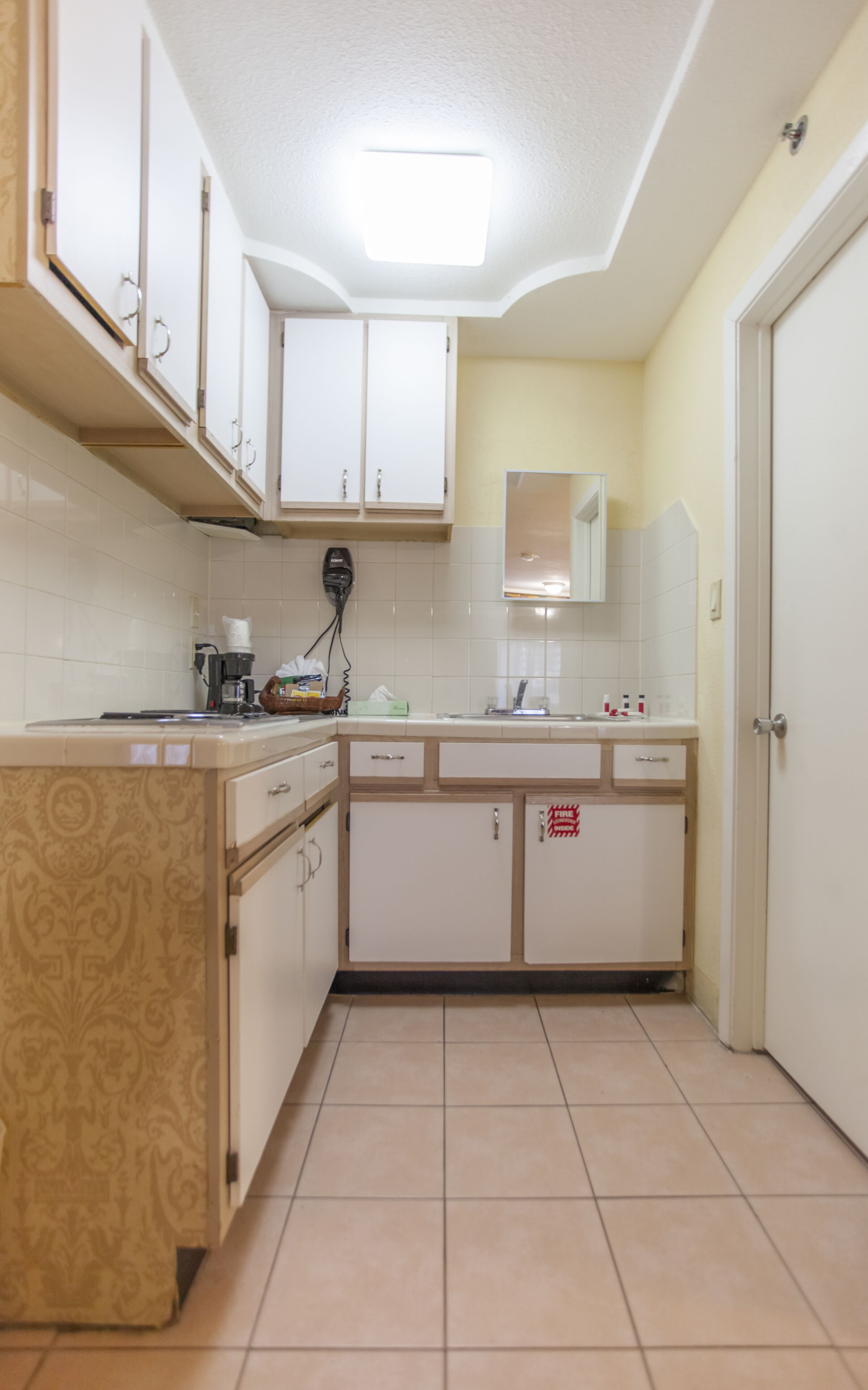 A kitchen with white cabinets, a tiled backsplash, and a tiled floor. There is a coffee maker on the counter and a fire extinguisher on the wall.