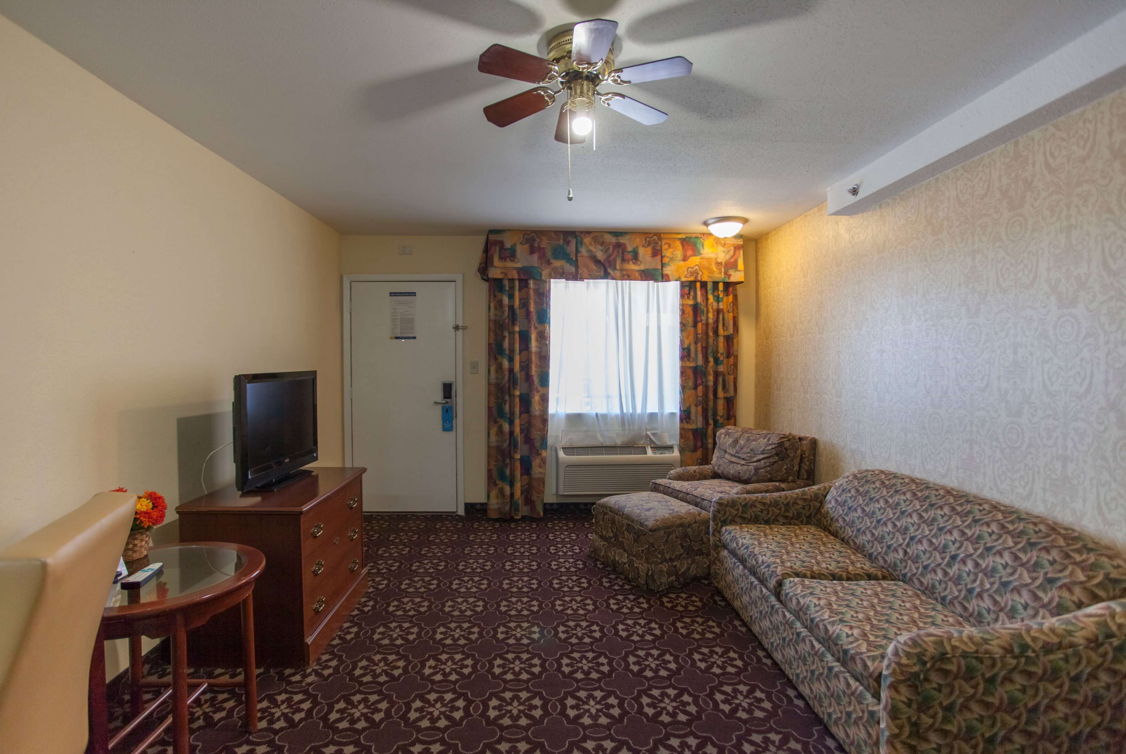 A hotel room with a ceiling fan, a television on a wooden dresser, a patterned sofa, a small table with a glass top, and a patterned carpet.