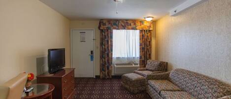 A hotel room with a ceiling fan, a television on a wooden dresser, a patterned sofa, a small table with a glass top, and a patterned carpet.