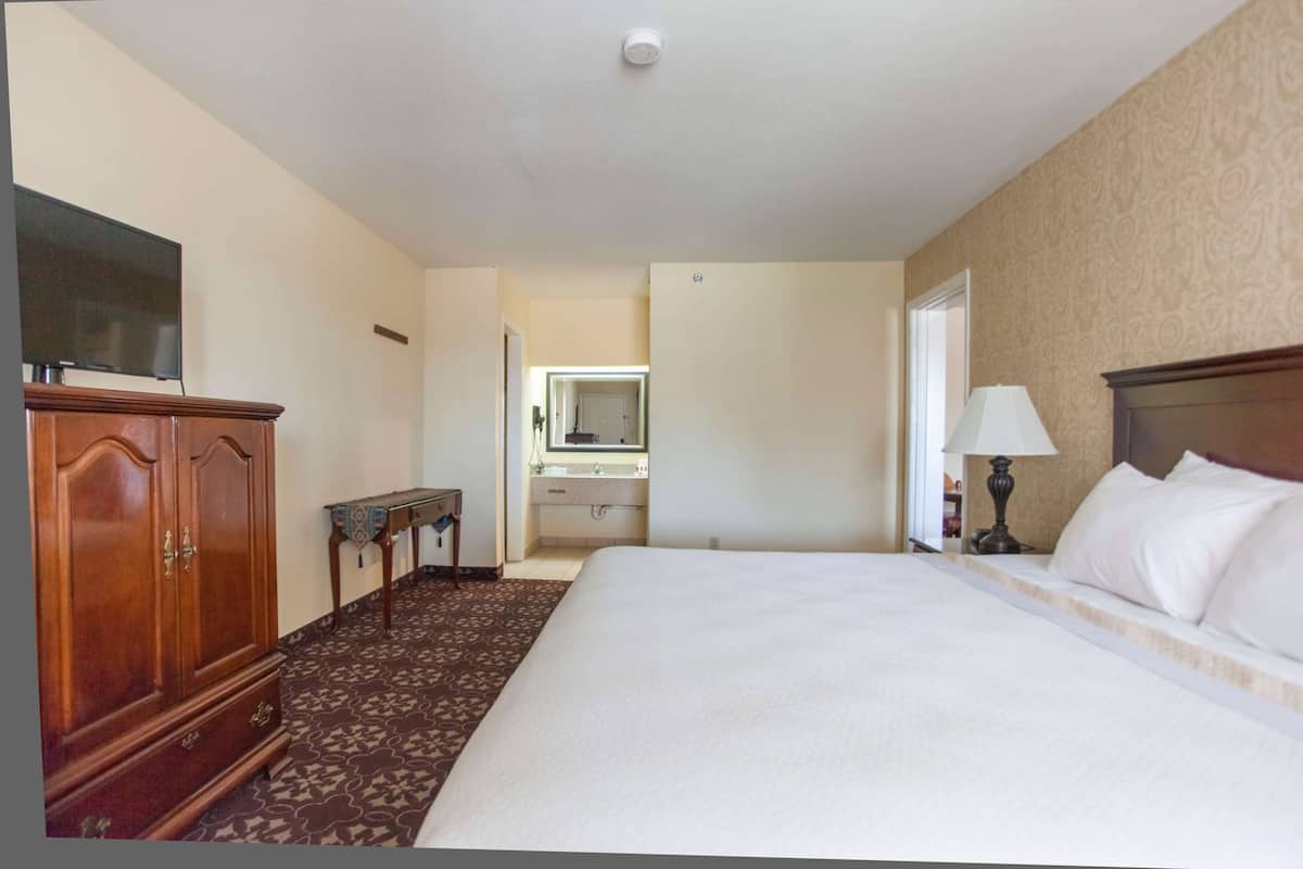 Une chambre d’hôtel avec un grand lit, une armoire en bois, un bureau avec un miroir et une salle de bain visible à travers une porte ouverte.