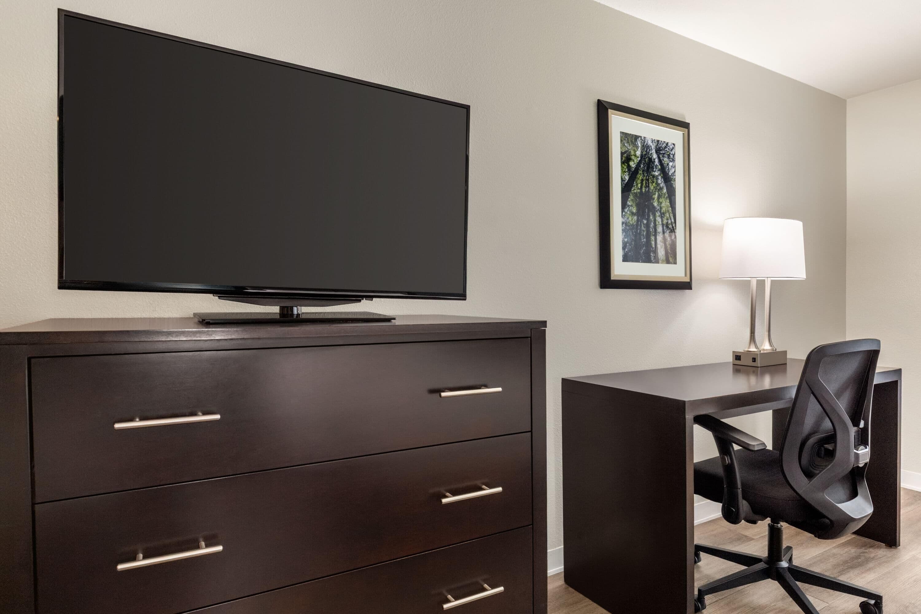 Habitación de hotel con televisor de pantalla plana sobre un mueble de madera oscura, un escritorio con una silla de oficina negra, un cuadro en la pared y una lámpara de mesa blanca.