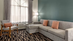 Ein modernes Hotelzimmer mit grauem Ecksofa, einem Holzschaukelstuhl, einem kleinen Tisch und einer Lampe.