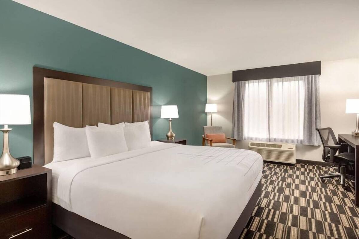Ein Hotelzimmer mit einem großen Bett, zwei Nachttischen mit Lampen, einem Schreibtisch mit Stuhl, einem Fenster mit Vorhängen und einem gemusterten Teppich.