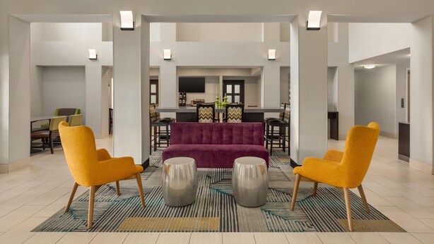 Eine geräumige Lobby mit einer zentralen Sitzecke, die ein violettes Sofa und zwei gelbe Sessel, einen gemusterten Teppich sowie eine moderne Küche im Hintergrund umfasst.