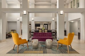 A spacious lobby with a central seating area featuring a purple sofa and two yellow chairs, a patterned rug, and a modern kitchen in the background.