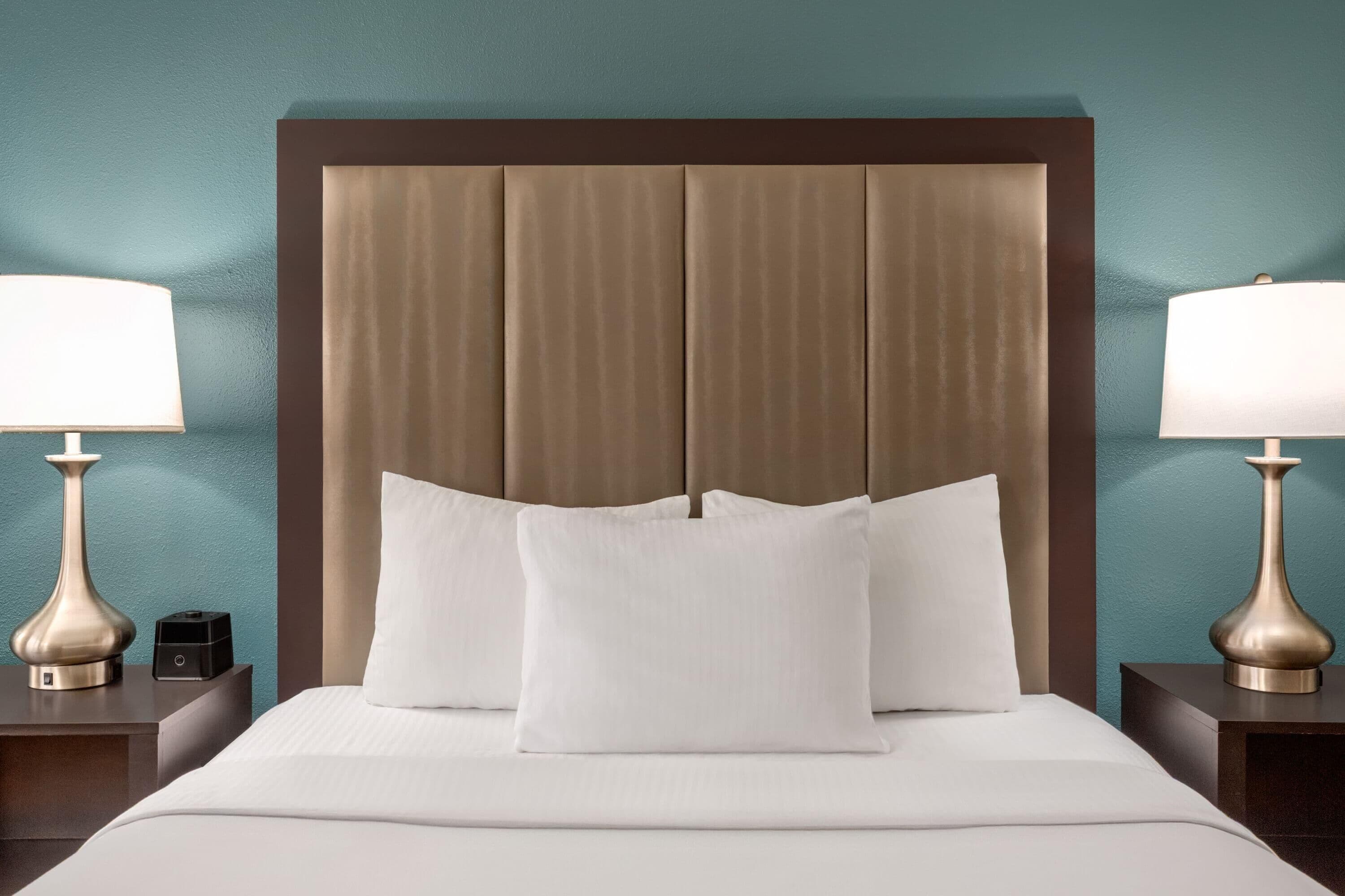 A neatly made bed with white linens and two pillows, flanked by two bedside tables with lamps and a small electronic device.