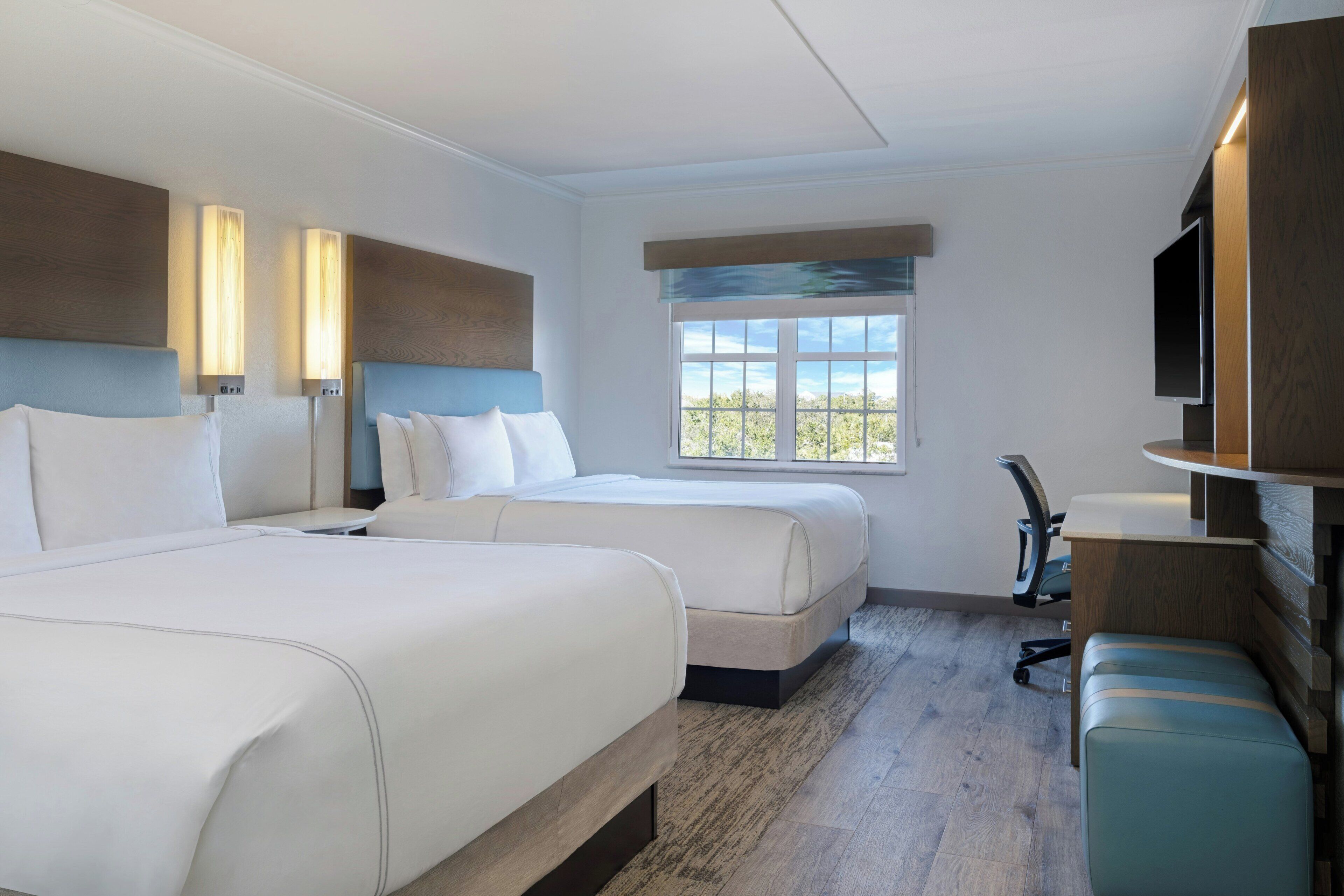 Quarto de hotel com duas camas, uma escrivaninha, uma cadeira, uma televisão e uma janela com vista.