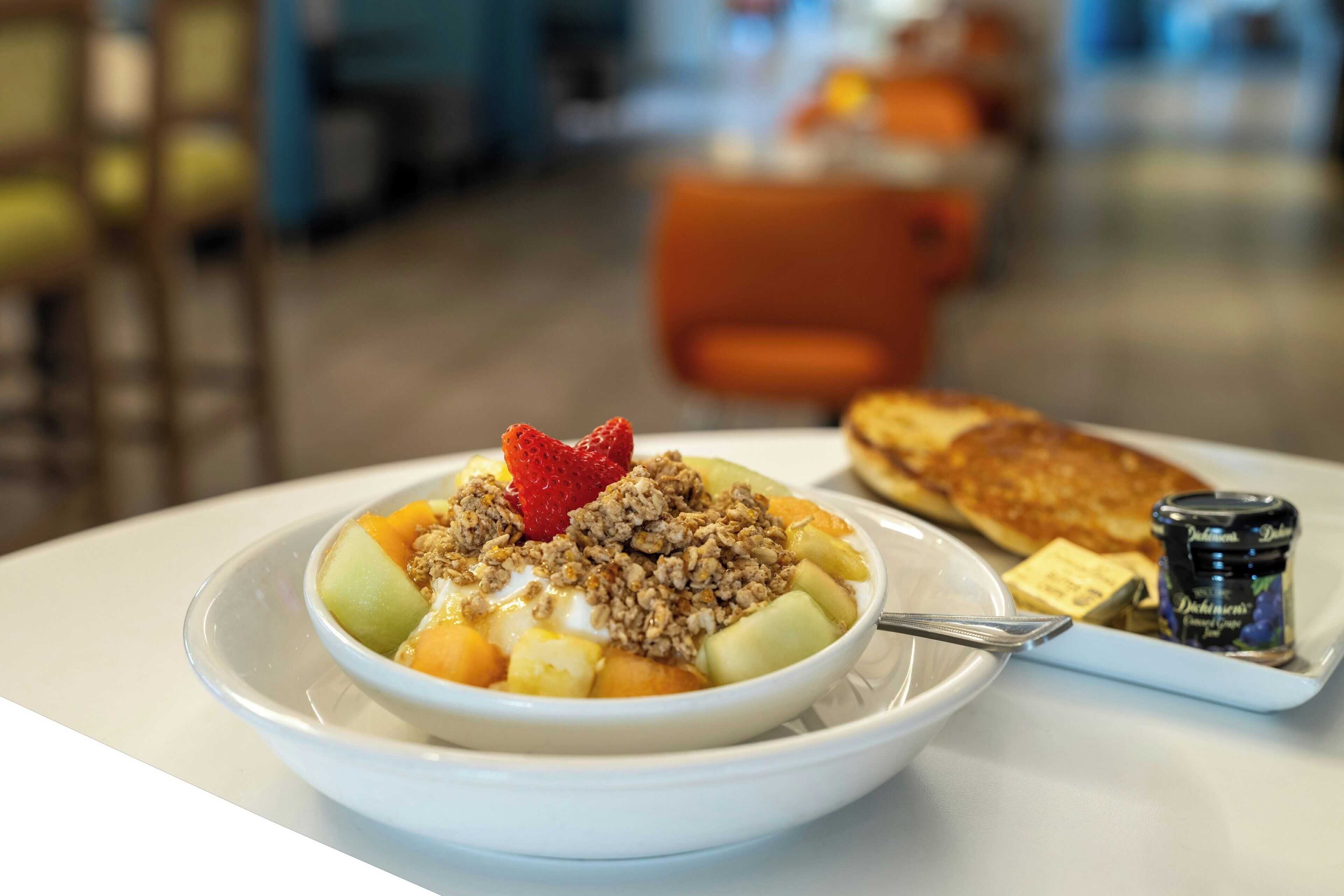 Desayuno con un tazón de fruta y granola, una porción de panqueques y un recipiente pequeño de jarabe.