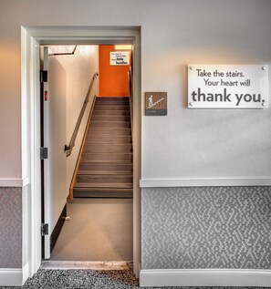 Un couloir avec une pancarte encourageant à utiliser les escaliers, une autre pancarte indiquant l’emplacement des escaliers, et un sol recouvert de moquette.
