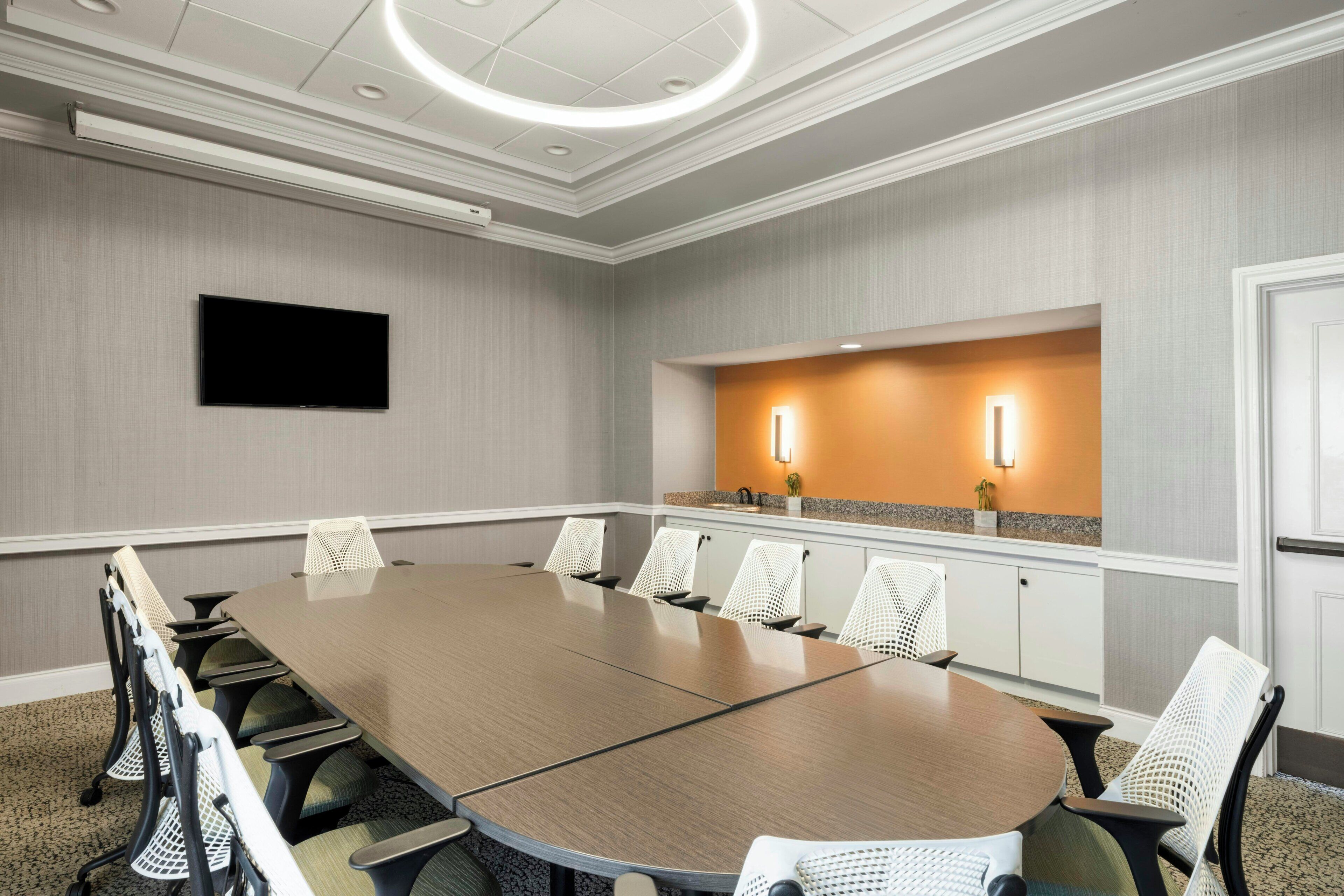 A conference room with a long table, white mesh chairs, a flat-screen TV, and two wall-mounted lights.