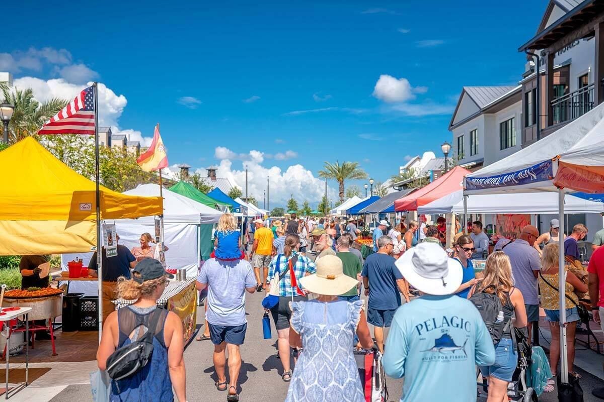 Un mercado al aire libre concurrido, con varios puestos y tiendas de campaña, gente caminando y un cielo azul despejado.