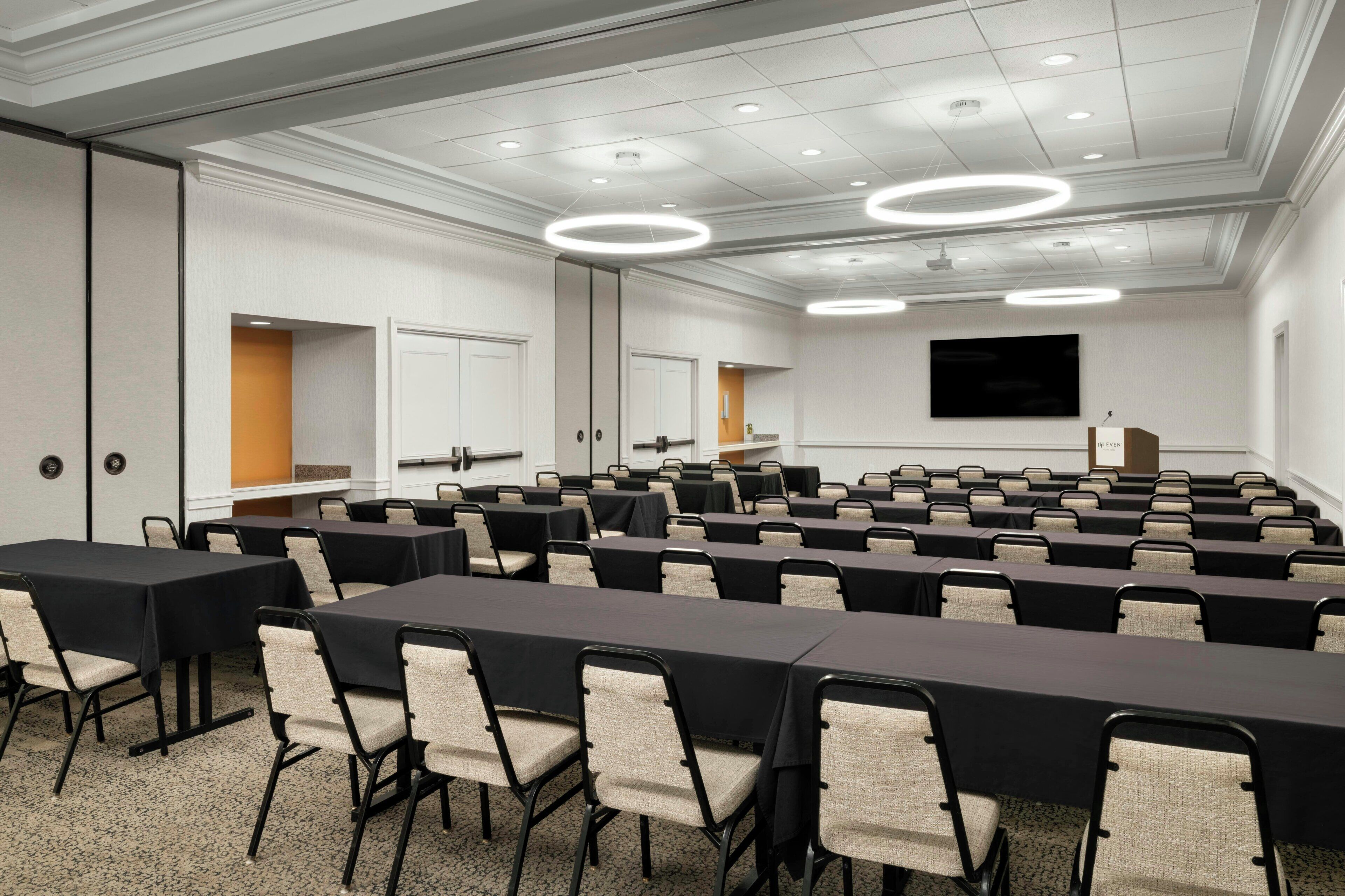 A conference room with rows of tables and chairs, a large screen, and a podium.