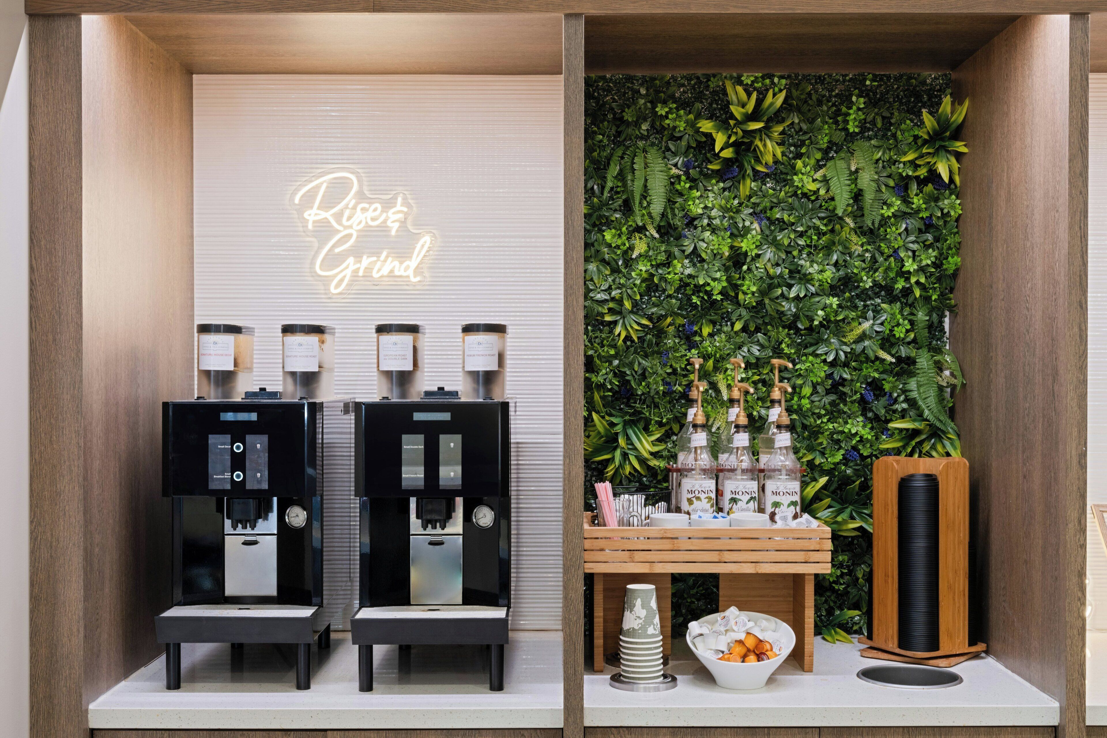 A coffee station with two espresso machines, a neon sign, and a wall of greenery.