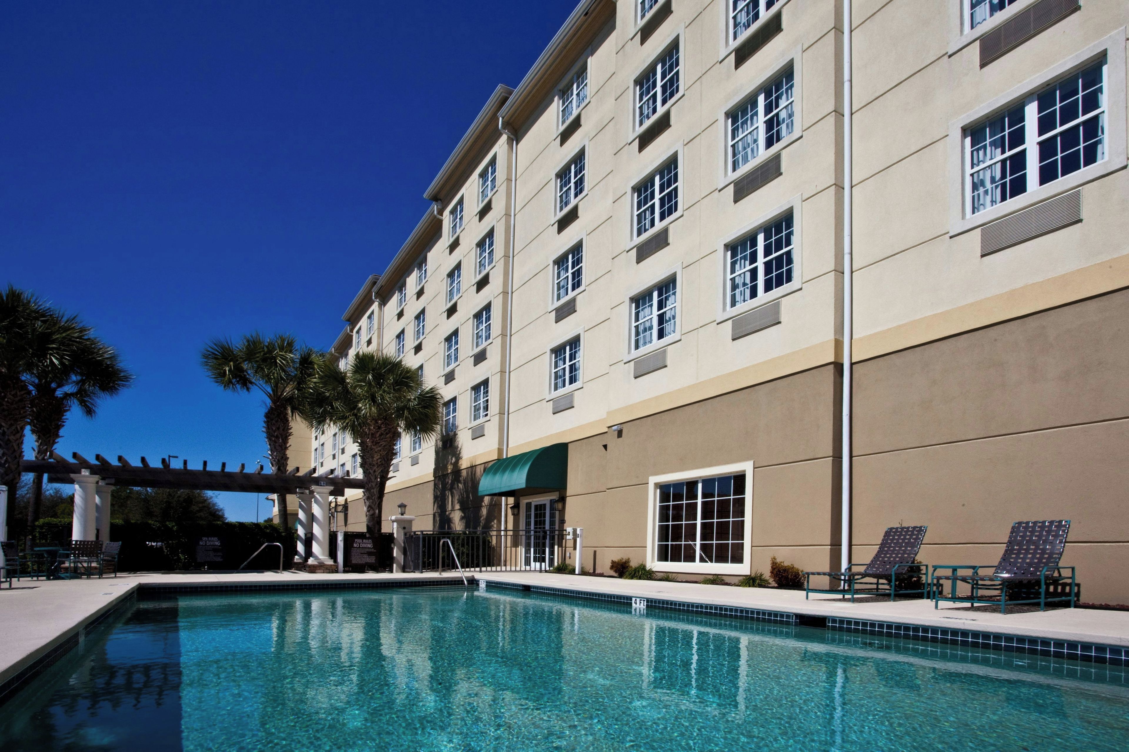 A hotel with a swimming pool, palm trees, and a covered area with chairs.
