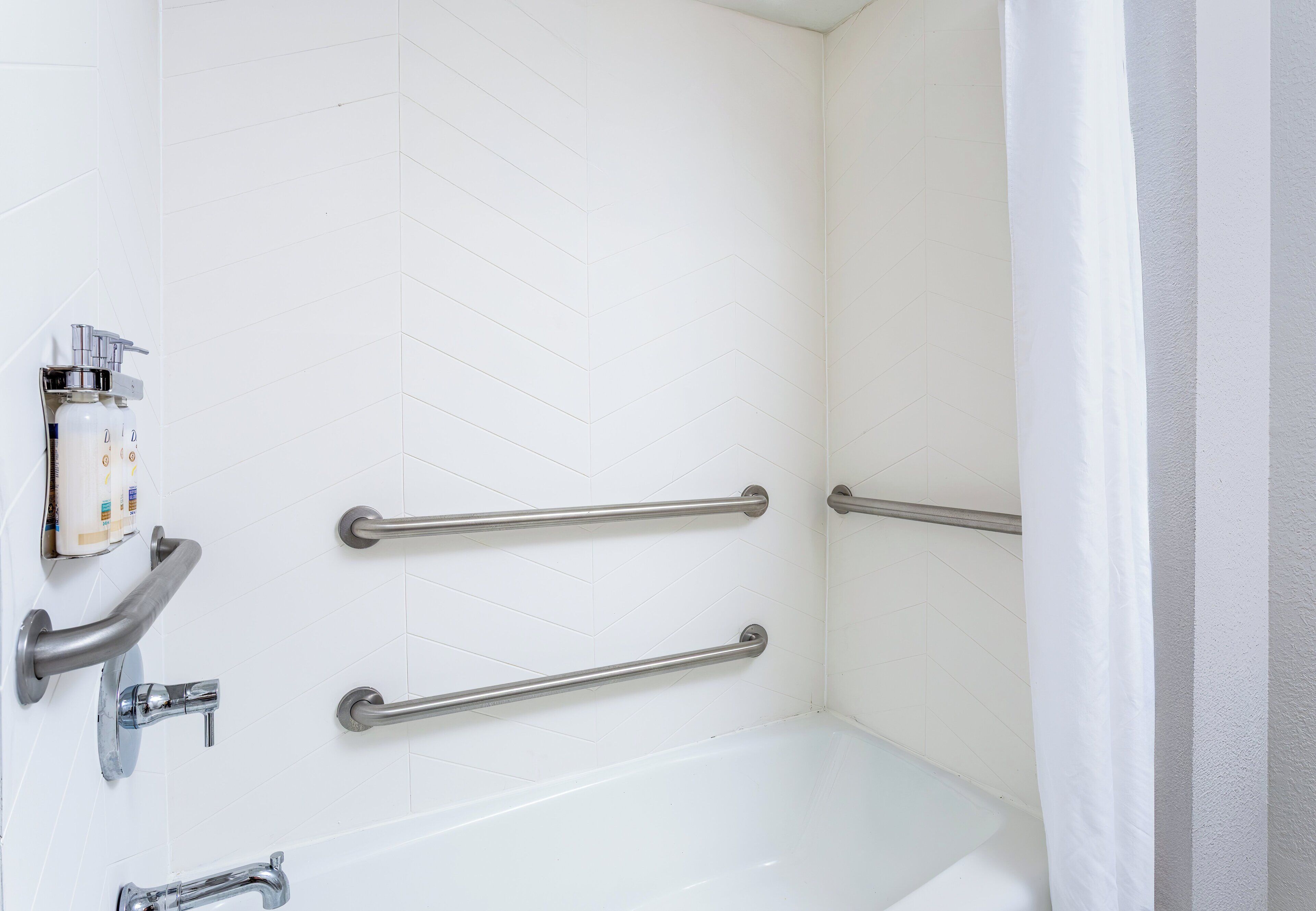 A bathroom with white tiled walls, a white bathtub, and two metal grab bars installed for support.