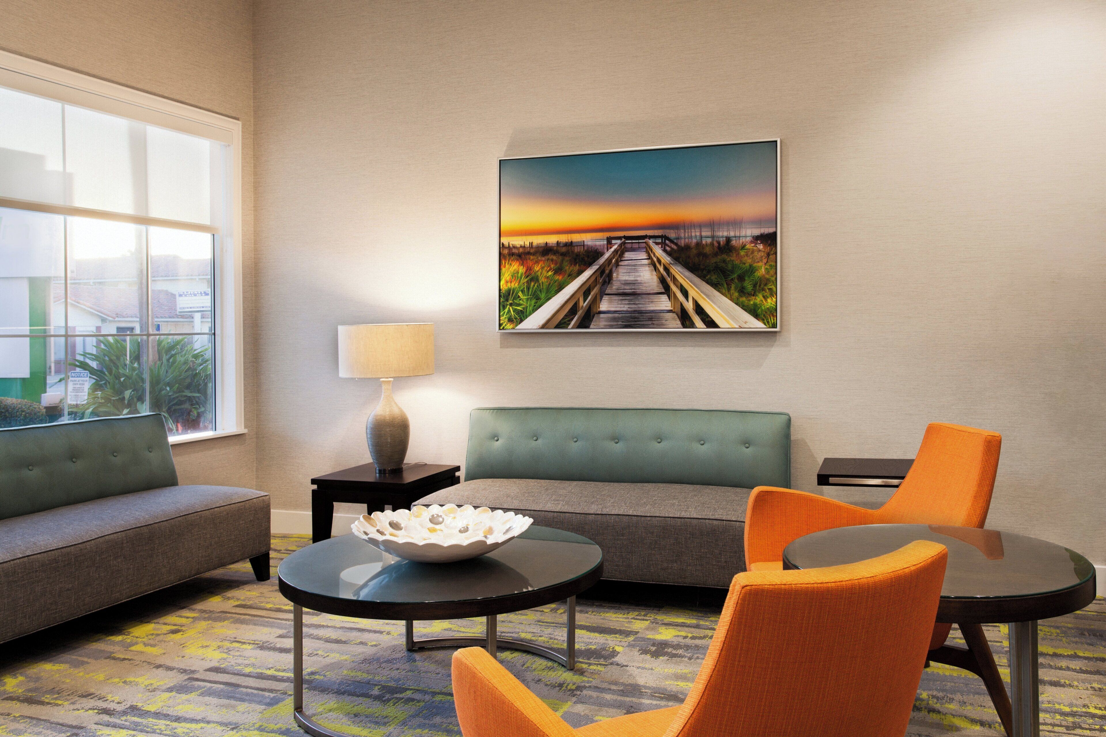 A hotel room with a sofa, two armchairs, a small table, a lamp, and a painting of a sunset over a bridge.