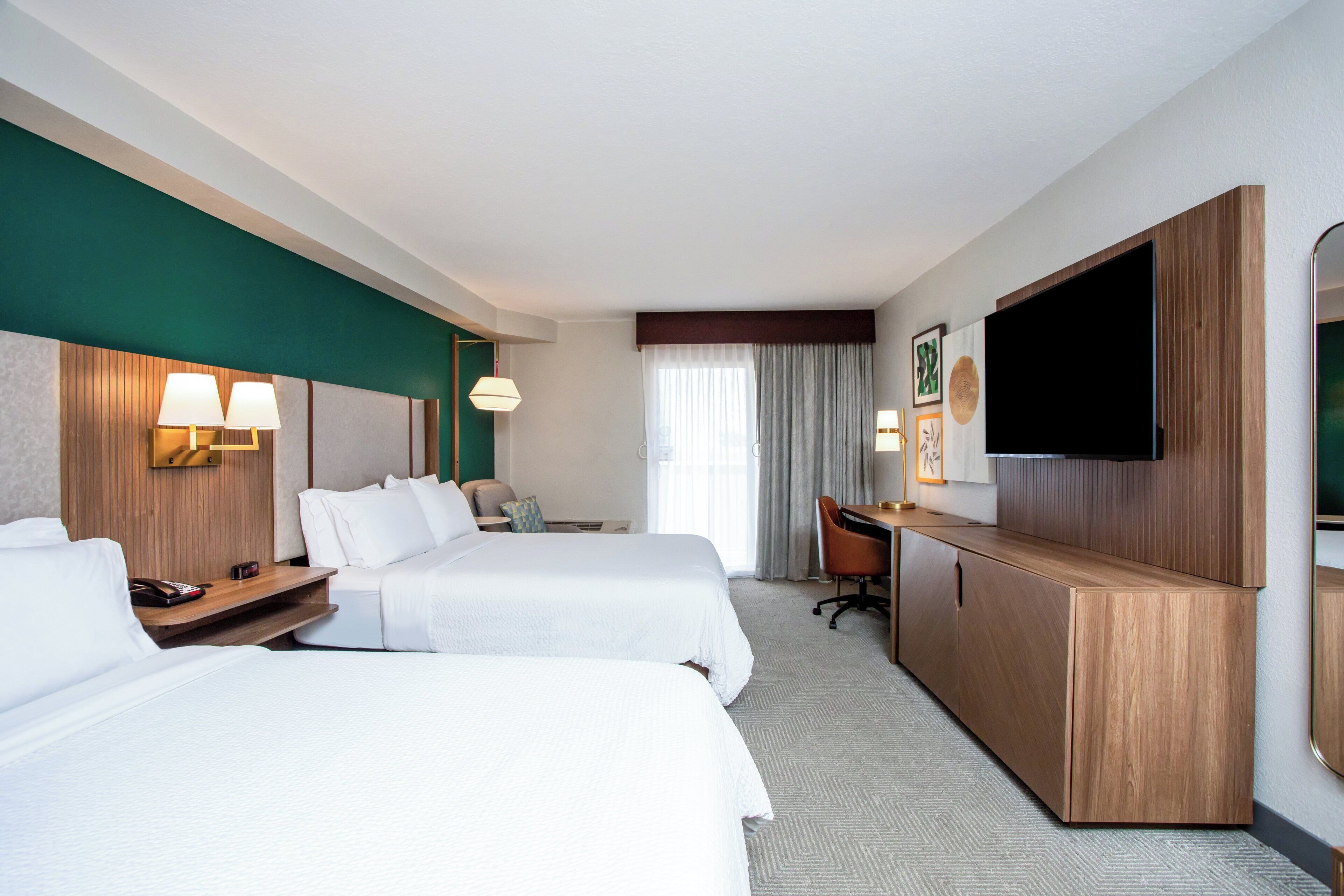 Une chambre d’hôtel avec deux lits, un bureau, une télévision et une grande fenêtre avec des rideaux.