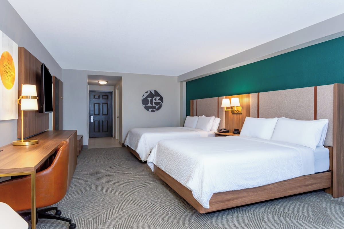 Een hotelkamer met twee bedden, een bureau en een stoel. Er is een televisie aan de muur en een deur die naar een andere kamer leidt.