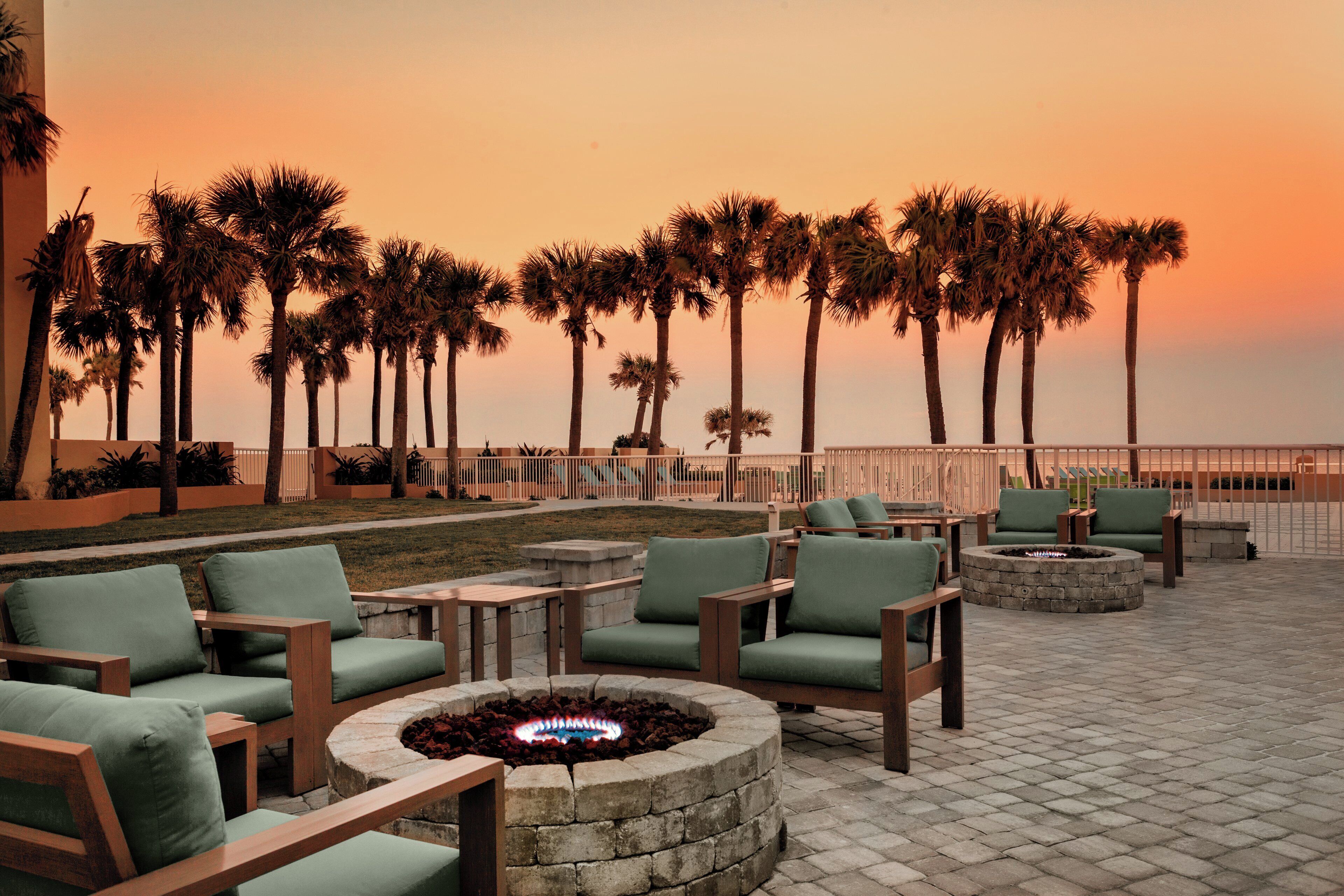 An outdoor seating area with fire pits and chairs, palm trees in the background, and a view of the ocean at sunset.