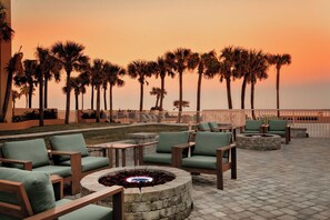 An outdoor seating area with fire pits and chairs, palm trees in the background, and a view of the ocean at sunset.