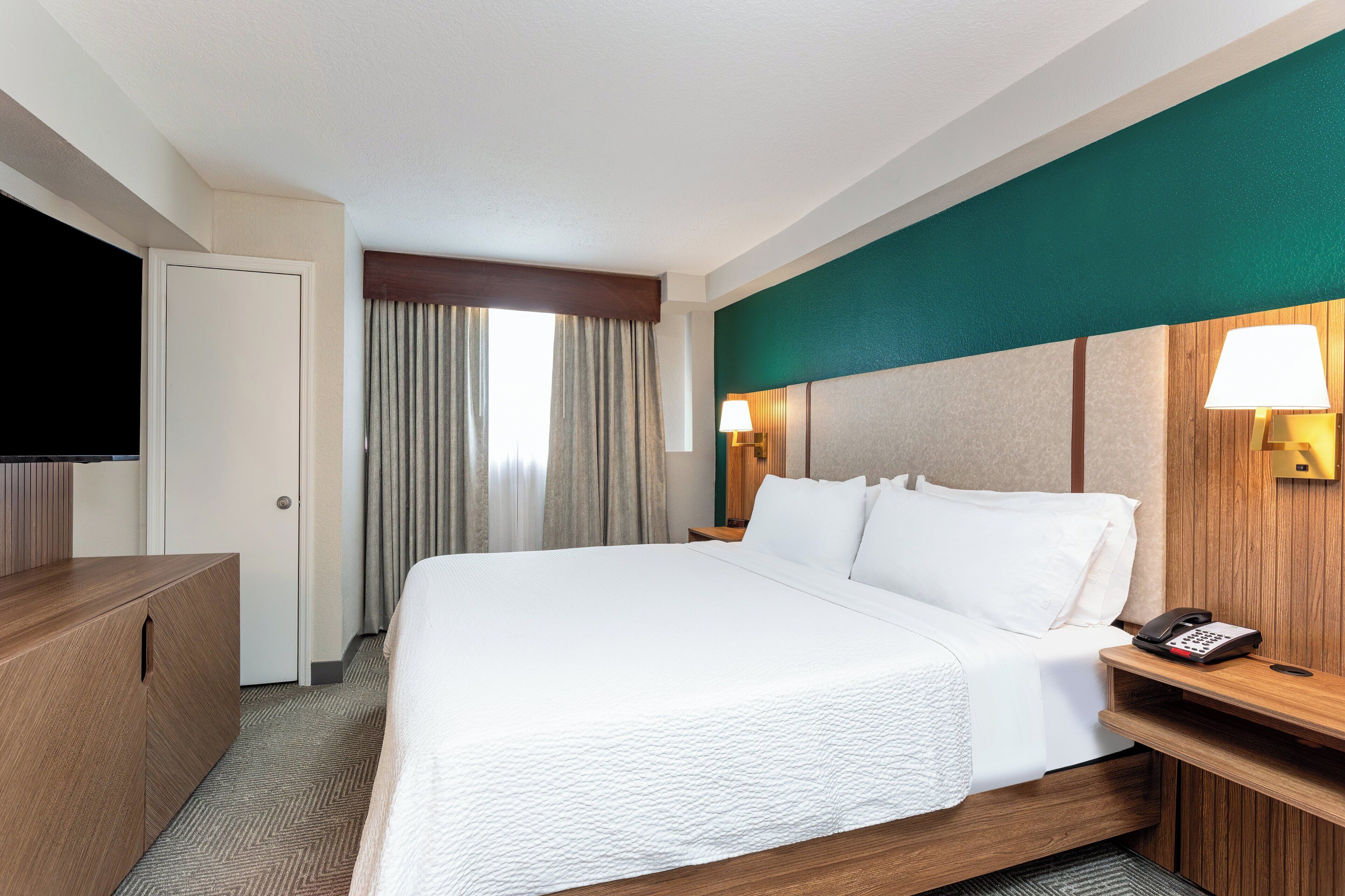 Una habitación de hotel con una cama grande, un escritorio de madera, un televisor y una lámpara fijada a la pared.