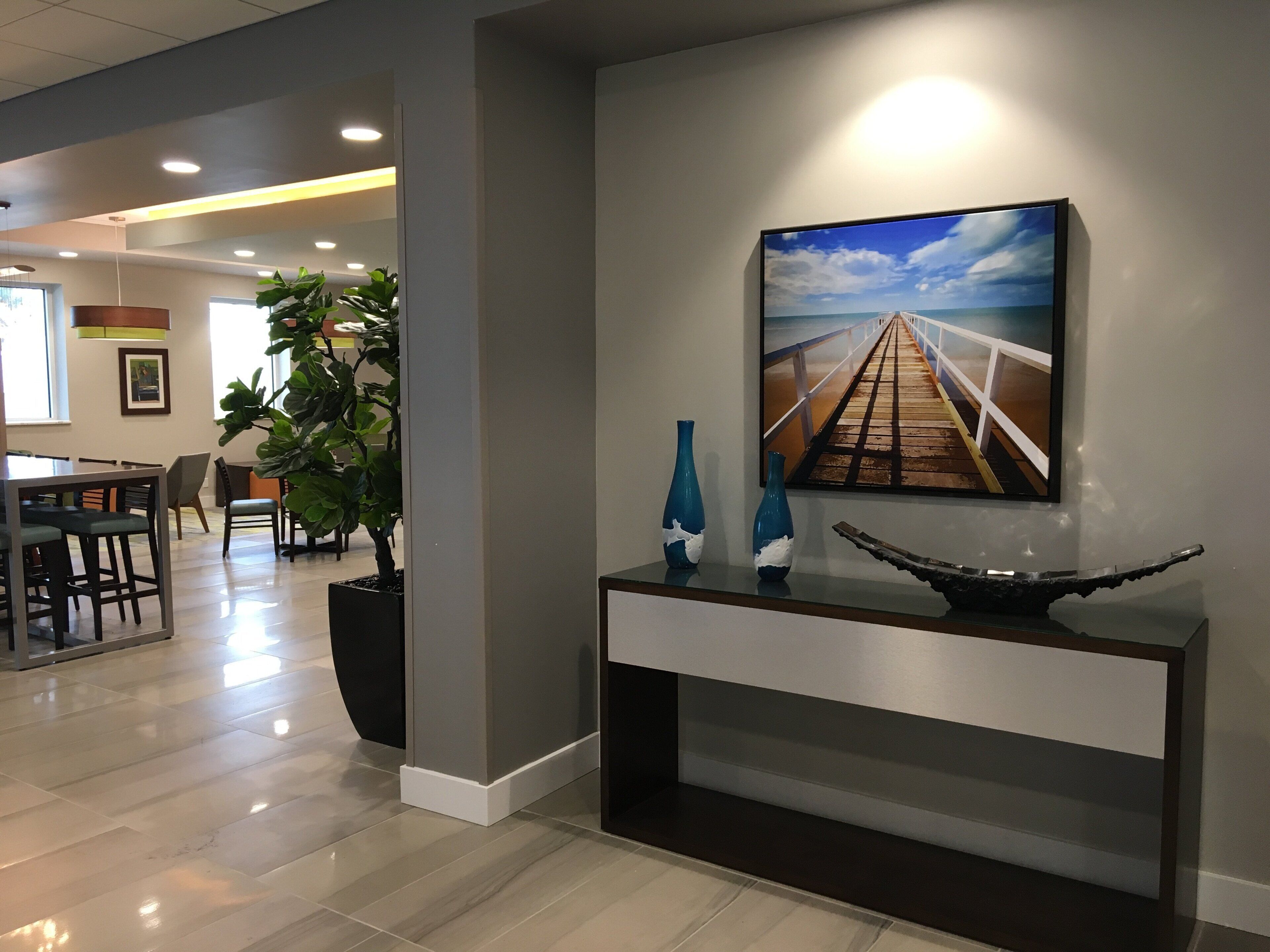A modern interior with a dining area, a large potted plant, a console table with decorative items, and a painting of a bridge.