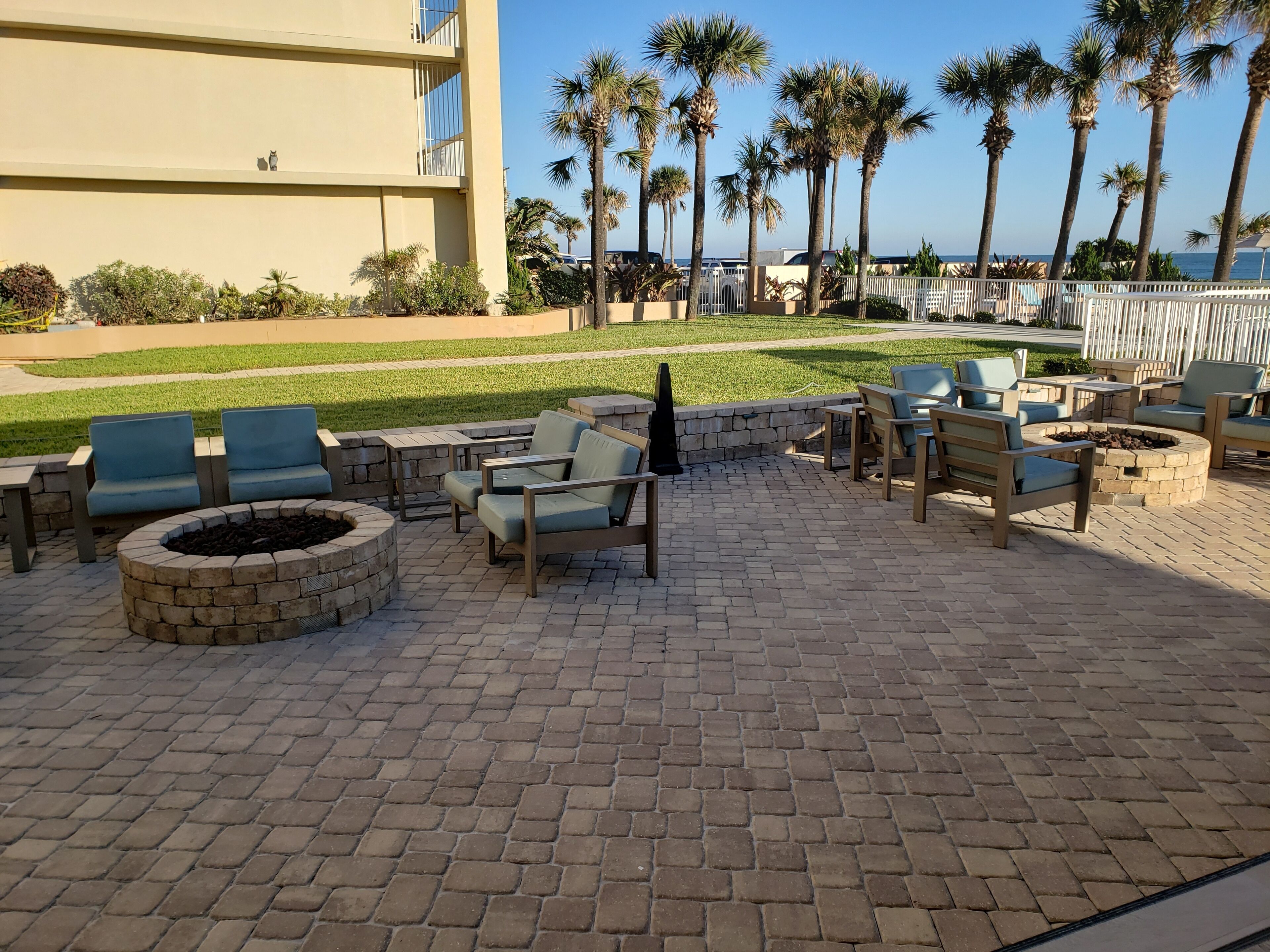 An outdoor seating area with fire pits and lounge chairs, surrounded by palm trees and a view of the ocean.