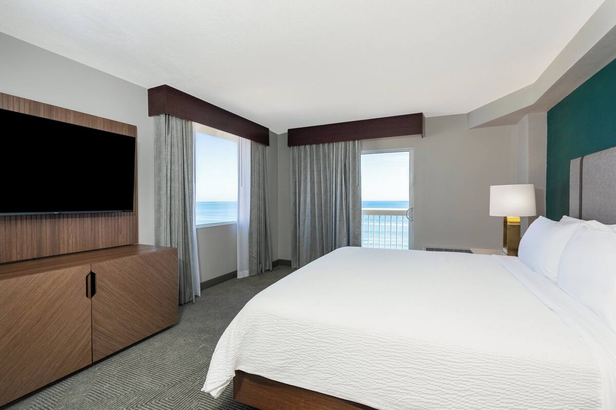 Een hotelkamer met een groot bed, een flatscreen televisie, een houten kast, een nachtkastje met een lamp, en uitzicht op de oceaan door de ramen.