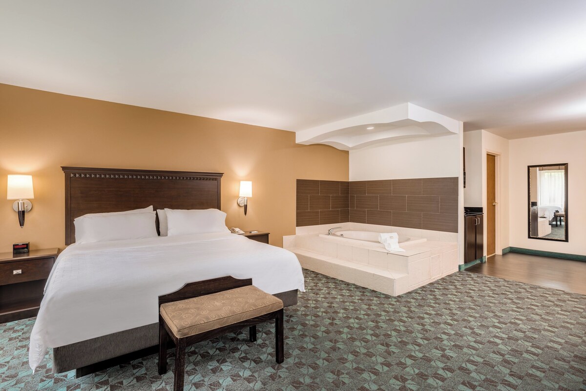 Una habitación de hotel con una cama grande, un banco, una bañera y un escritorio pequeño con una lámpara.