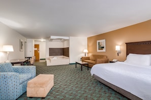 Habitación de hotel con una cama grande, un sofá, una silla, una mesita, una lámpara, una mesita de noche y un baño con bañera y ducha.