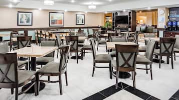 Ein Speisebereich eines Hotels mit mehreren Tischen und Stühlen, einem Flachbildfernseher und einer Kaffeestation.