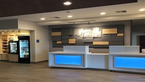 A reception area with a modern design, featuring a counter with illuminated blue panels, a wall-mounted sign, and a refrigerator stocked with beverages. - Holiday Inn Express & Suites Albany by IHG (Albany)