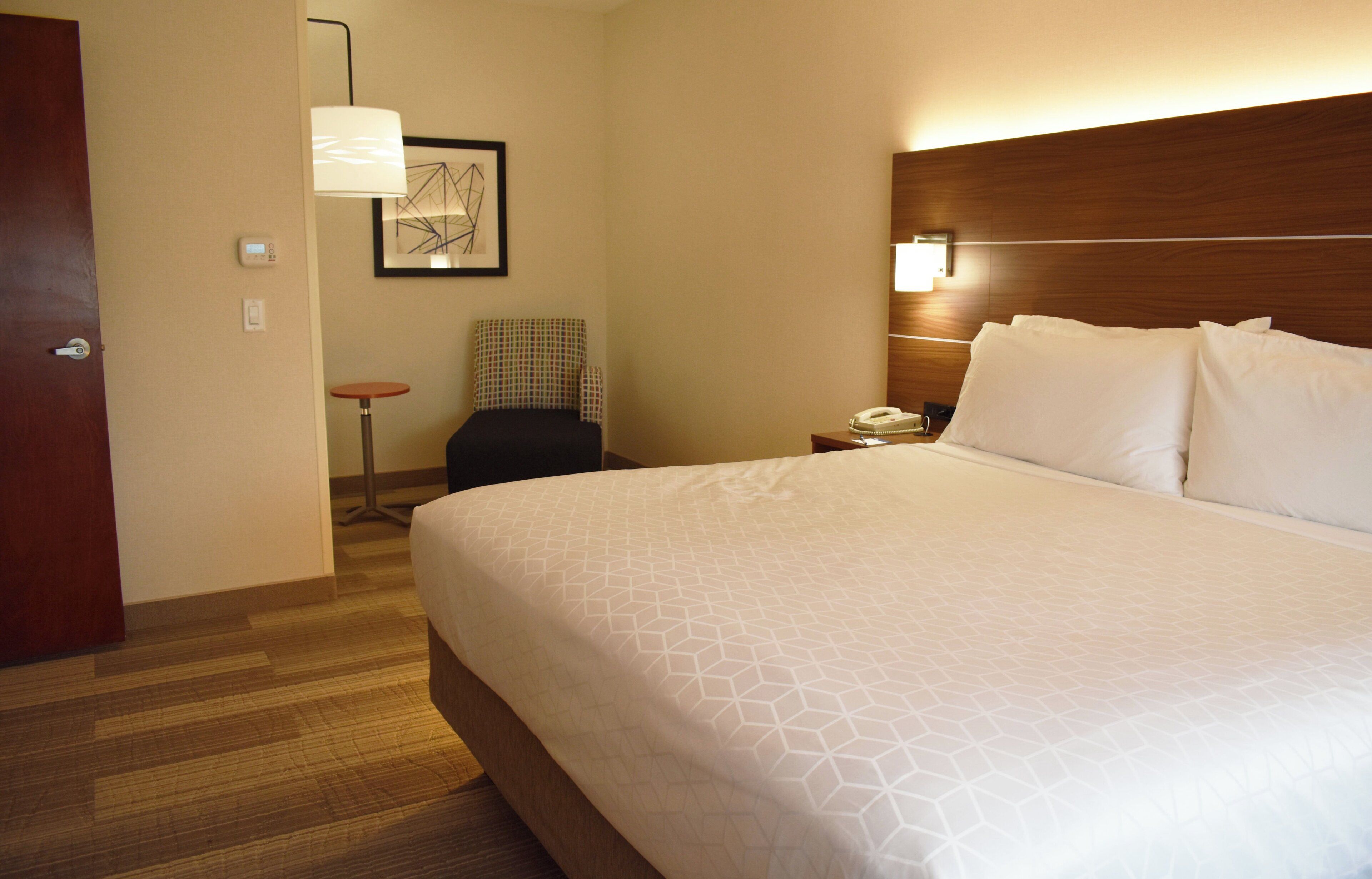 Habitación de hotel con una cama grande, una mesita de noche con una lámpara, una silla y una pequeña mesa con un teléfono y una taza.