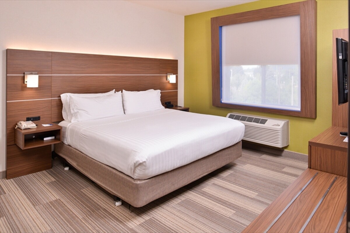 A hotel room with a large bed, bedside tables, a wall-mounted TV, and a window with blinds.