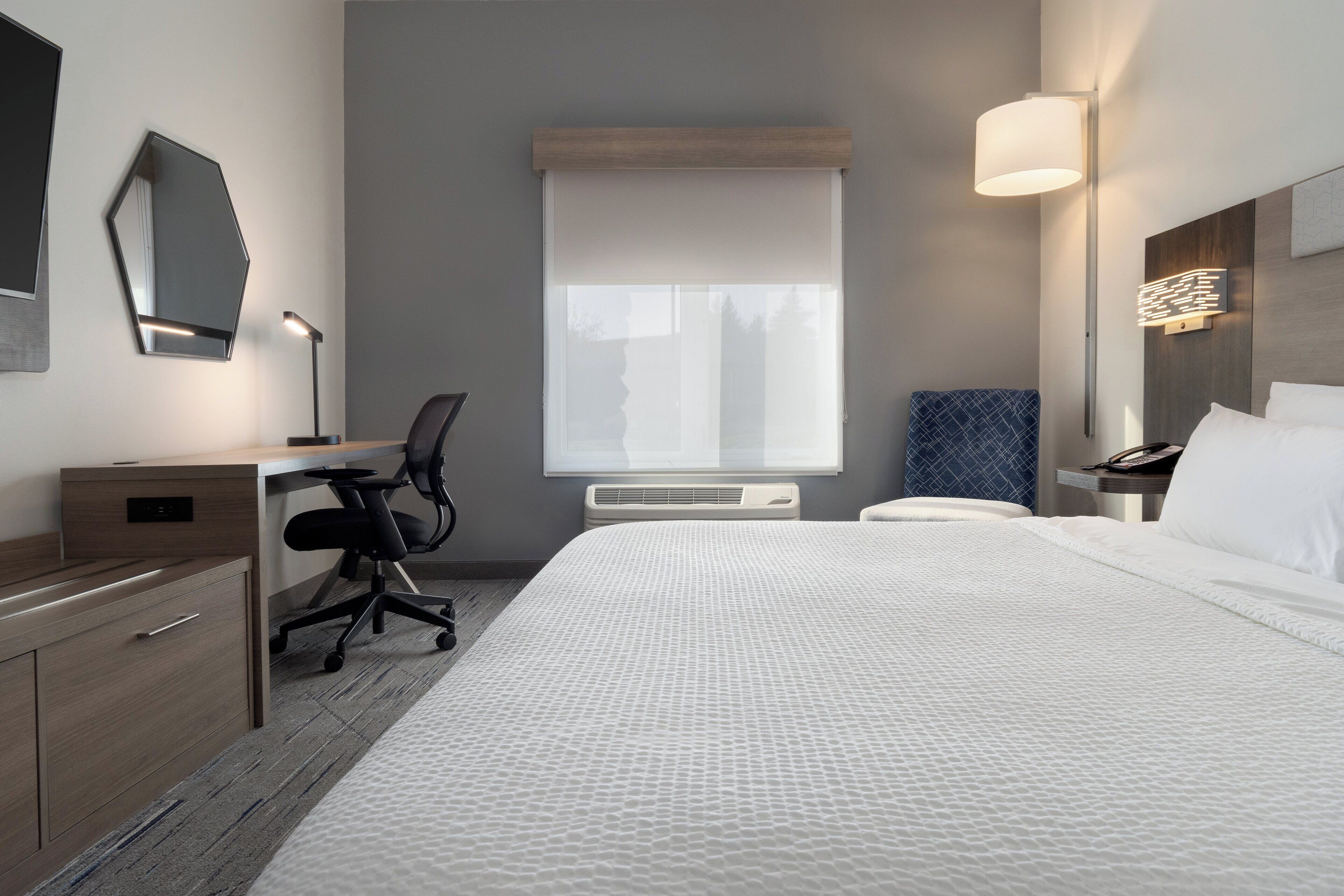 Habitación de hotel con una cama grande, un escritorio con silla, una mesita de noche y una lámpara de pared.