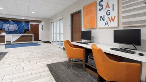 A modern office space with two desks, each with a computer and an orange chair. There are framed artworks on the wall and a reception area in the background. - Holiday Inn Express Hotel & Suites Saginaw by IHG (Saginaw)