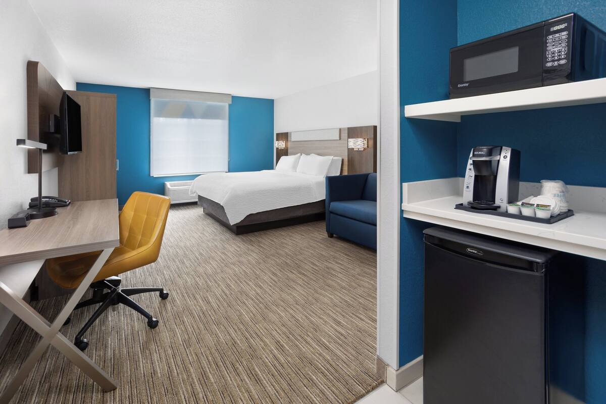 A hotel room with a bed, a desk with a chair, a microwave, a coffee maker, and a television.