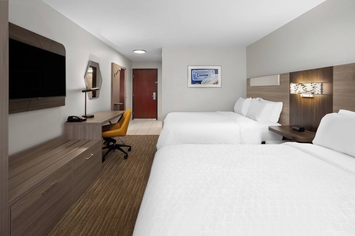 A hotel room with two beds, a desk, a chair, a TV, and a wall-mounted light fixture.