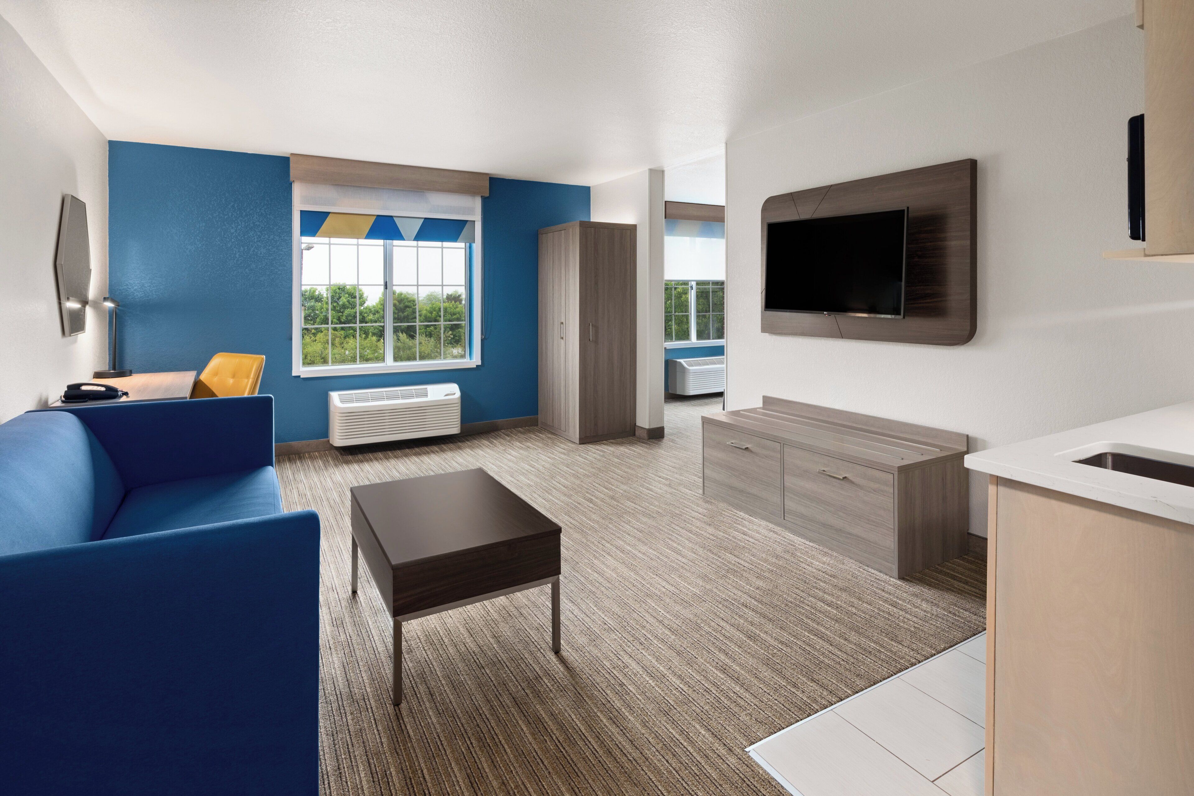 A modern hotel room with a blue sofa, a small table, a TV mounted on the wall, and a kitchenette area.