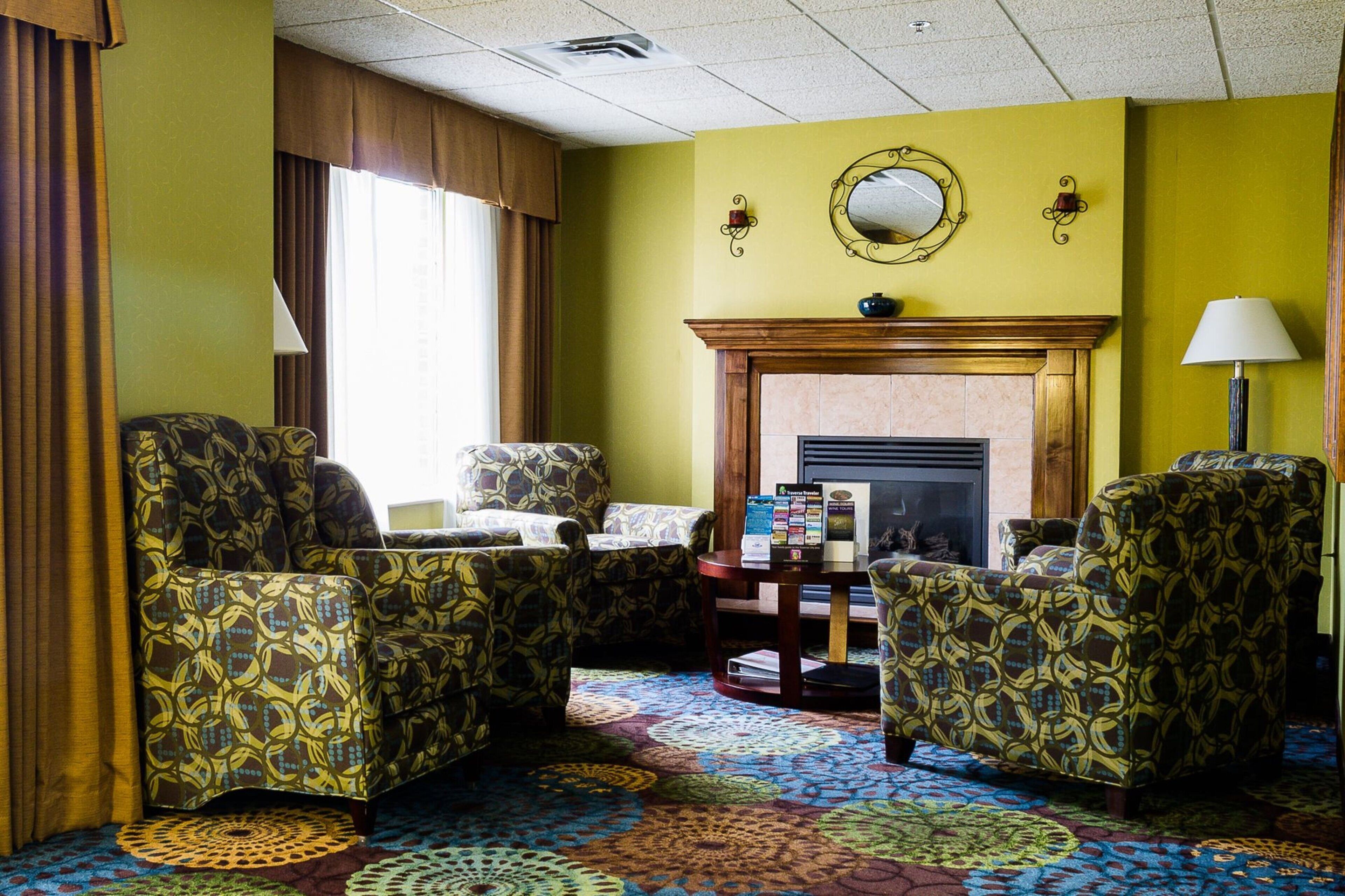 A hotel room with a fireplace, patterned armchairs, a round coffee table, and a colorful carpet.