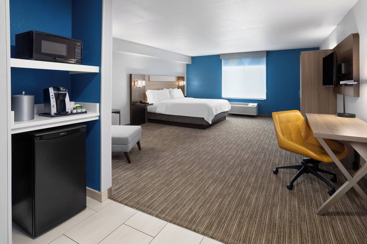 A hotel room with a bed, desk, chair, microwave, coffee maker, and a TV.