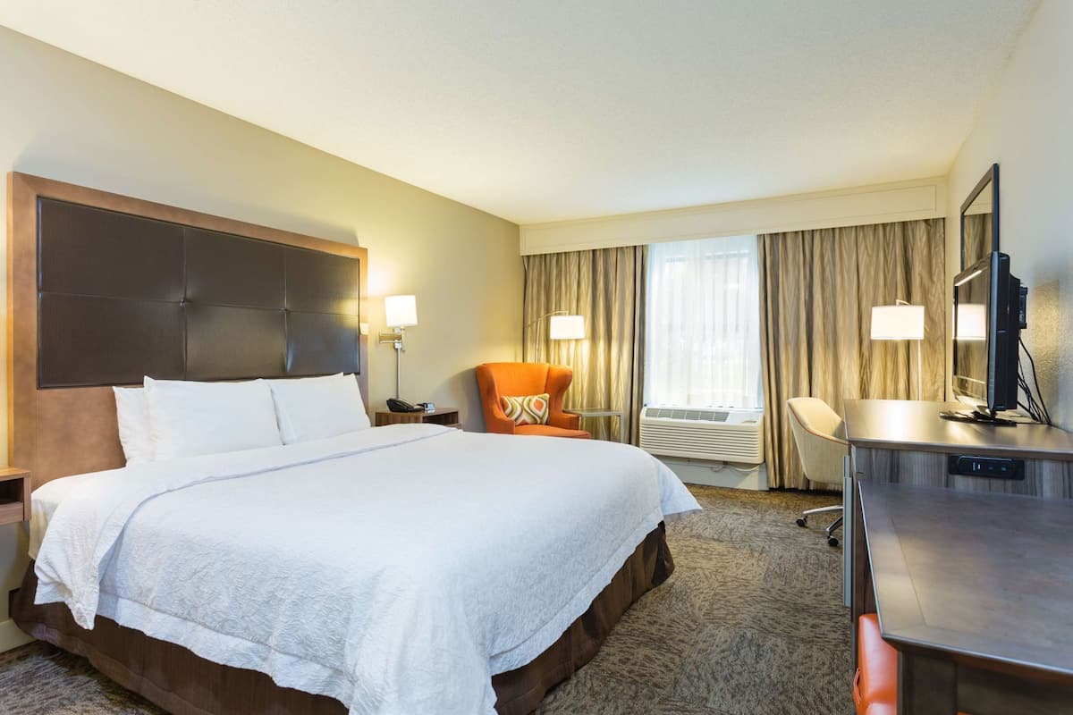 Habitación de hotel con una cama grande, un escritorio con computadora, una silla, una lámpara y una ventana con cortinas.