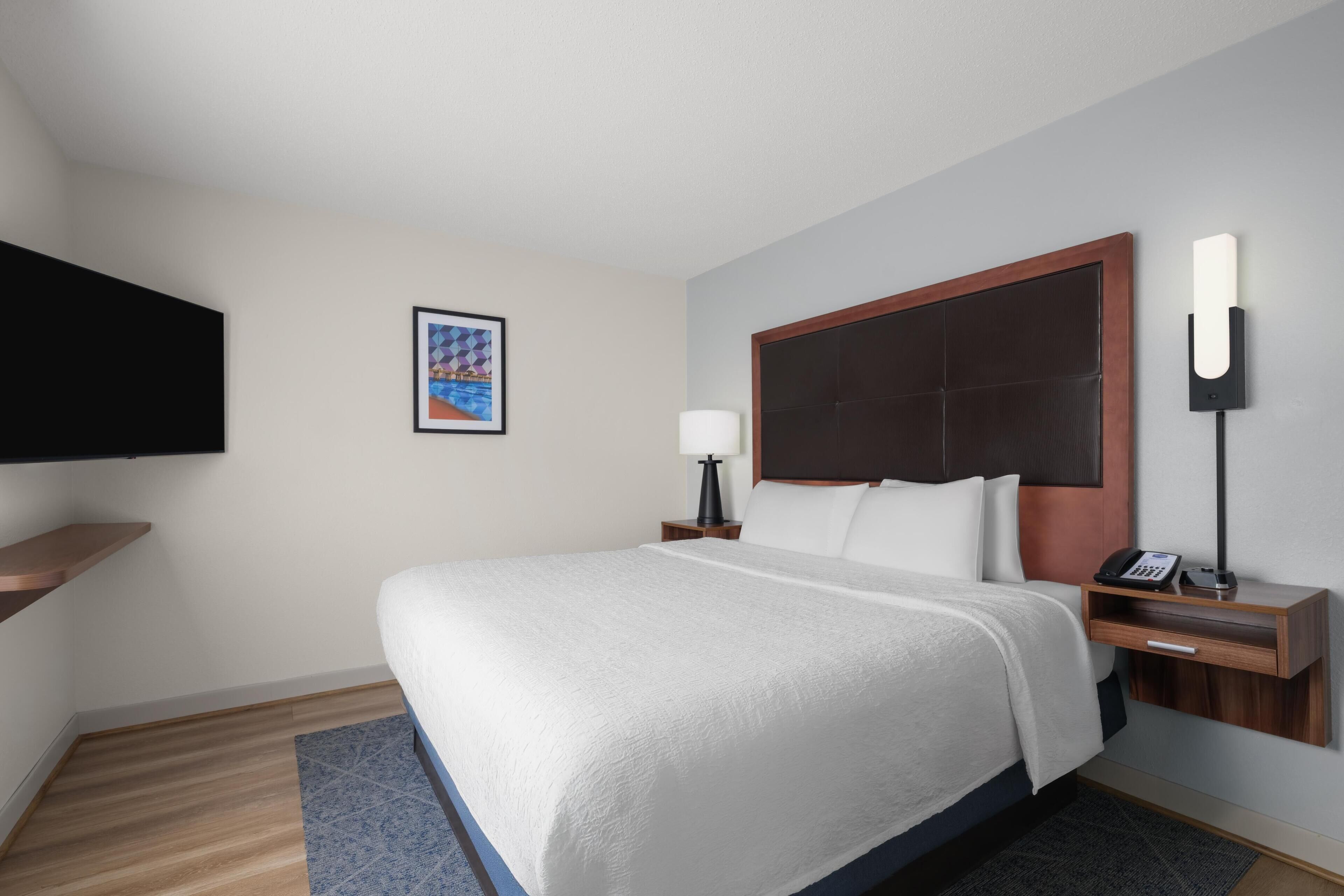 Une chambre d’hôtel avec un grand lit, une table de chevet avec une lampe, un téléviseur à écran plat fixé au mur et un tableau encadré.