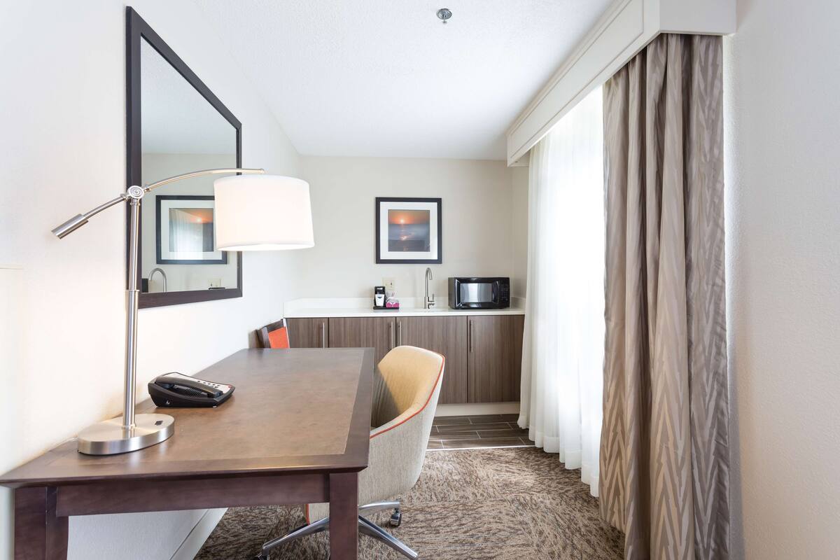 Habitación de hotel con escritorio, silla, lámpara, microondas y vistas al exterior por la ventana.