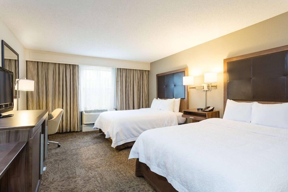 A hotel room with two beds, a desk, a chair, a TV, and a window with curtains.
