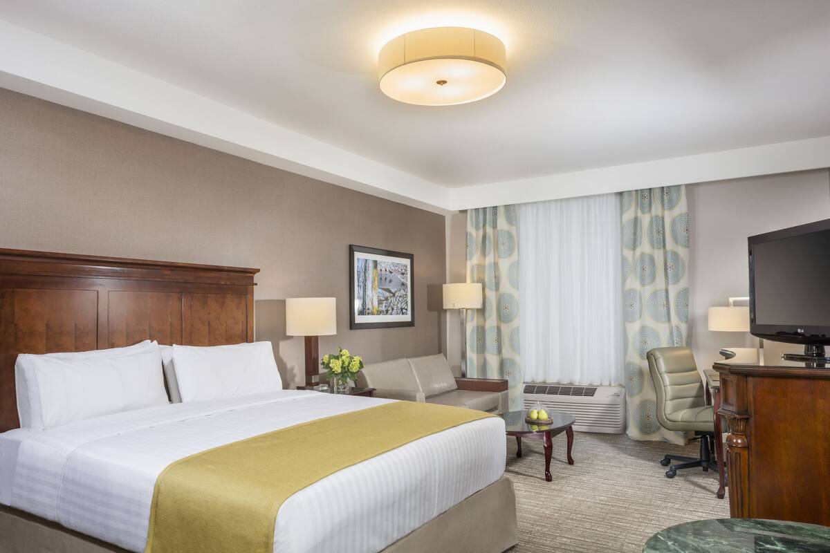 Una habitación de hotel con una cama grande, dos mesitas de noche con lámparas, un escritorio con silla, un televisor y un ventanal con cortinas estampadas.
