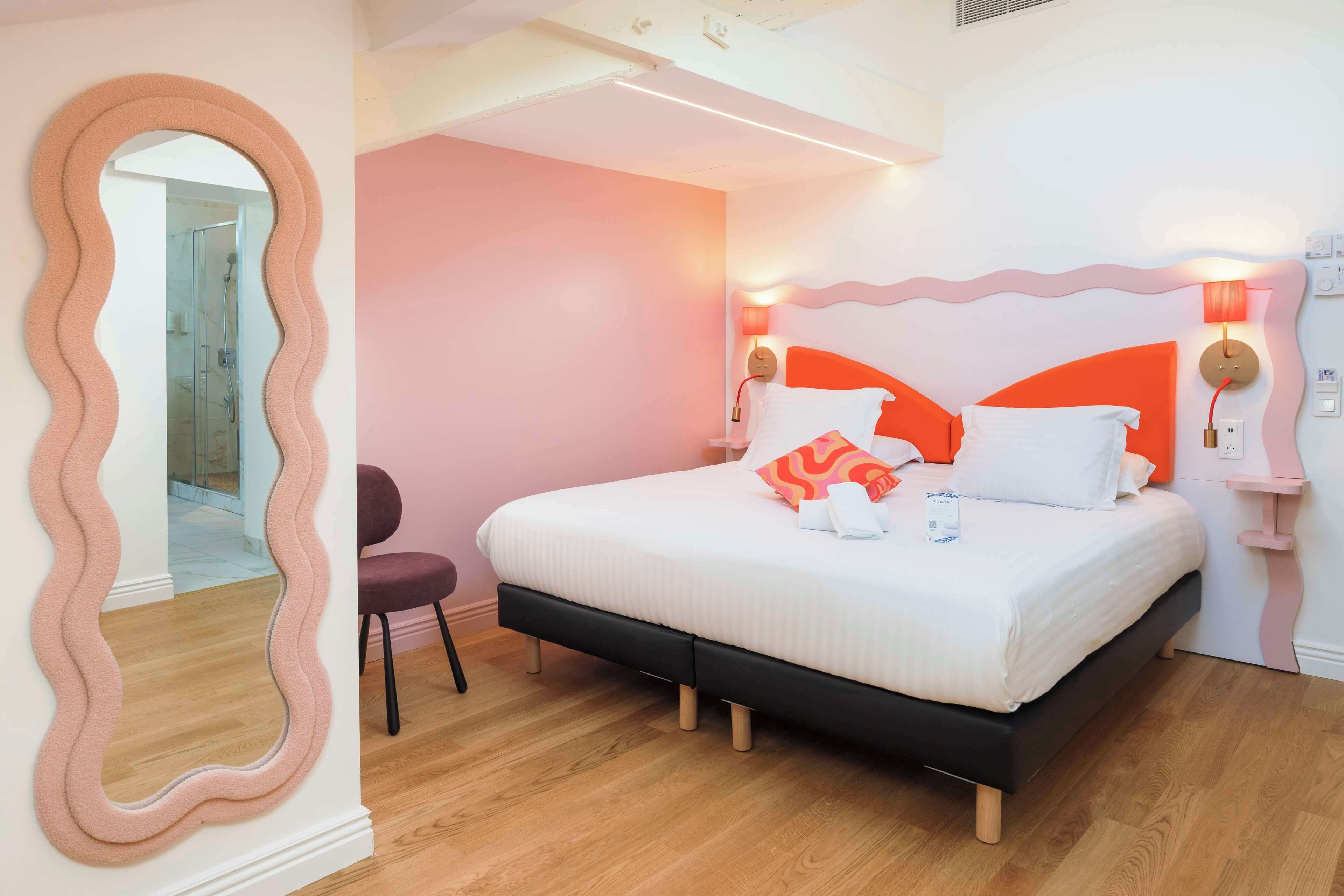 A bedroom with a bed, a chair, a mirror, and a bathroom visible through an archway.