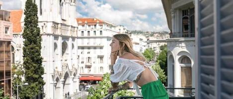 Uma pessoa apoiada na balaustrada de uma varanda, com uma paisagem urbana ao fundo.