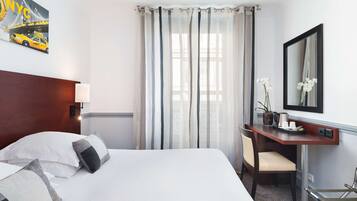 Habitación de hotel con una cama grande, un escritorio con silla, un espejo y una ventana con cortinas translúcidas.